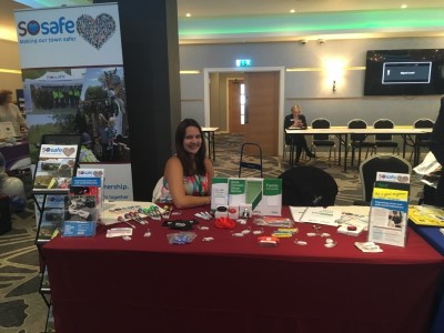 Jessica sits at a table at an exhibition.  The table displays a range of promotional material for the Stevenage Safety team, such as pens, key rings and badges